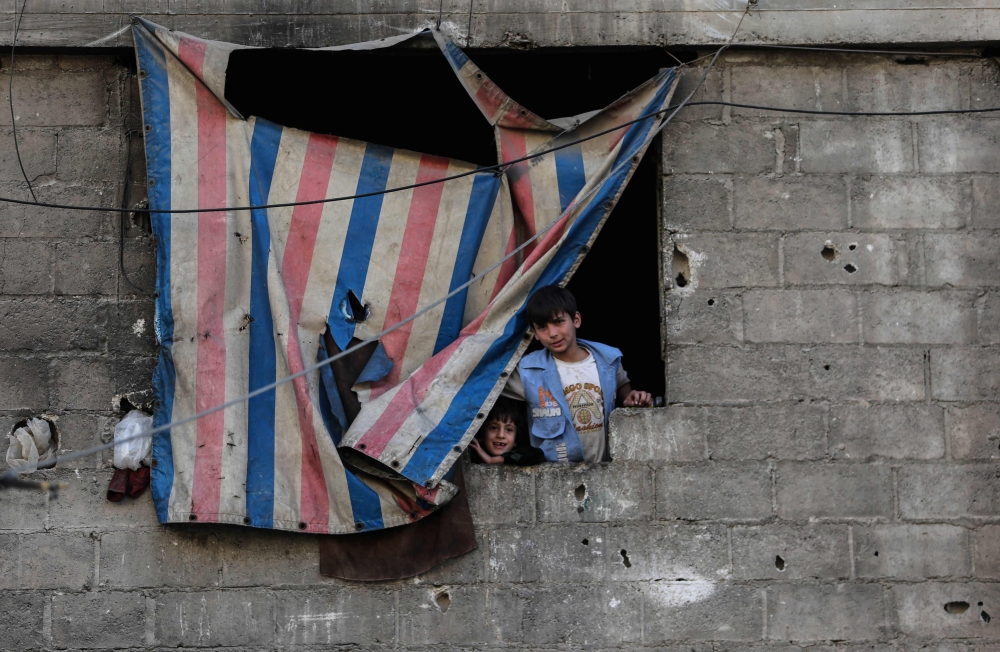 TOPSHOT - Syrian boys stand at a window covered with a makeshift curtain in Douma, a rebel-held town on the outskirts of the capital Damascus, on October 24, 2017. According to the World Bank, the conflict in Syria has cost the country's economy some $226 billion -- about four times the country's gross domestic product in 2010. The fighting has damaged or destroyed 27 percent of Syria's housing stock and about half the country's medical and educational facilities. About 85 percent of the population lives below the poverty line and half are unemployed. 
 / AFP / Amer ALMOHIBANY
