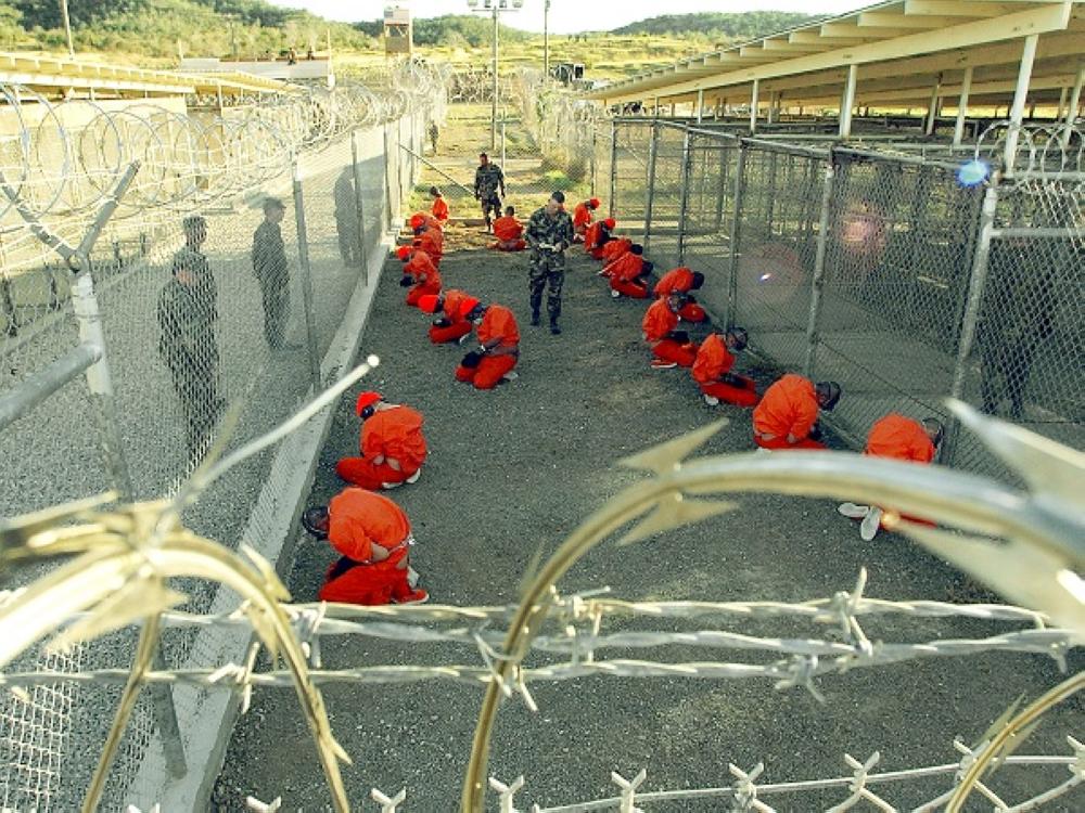 GUANTANAMO BAY, CUBA - JANUARY 11: In this handout photo provided by the U.S. Navy, U.S. Military Police guard Taliban and al Qaeda detainees in orange jumpsuits January 11, 2002 in a holding area at Camp X-Ray at Naval Base Guantanamo Bay, Cuba during in-processing to the temporary detention facility. The detainees will be given a basic physical exam by a doctor, to include a chest x-ray and blood samples drawn to assess their health, the military said. The U.S. Department of Defense released the photo January 18, 2002. (Photo by Petty Officer 1st class Shane T. McCoy/U.S. Navy via Getty Images)