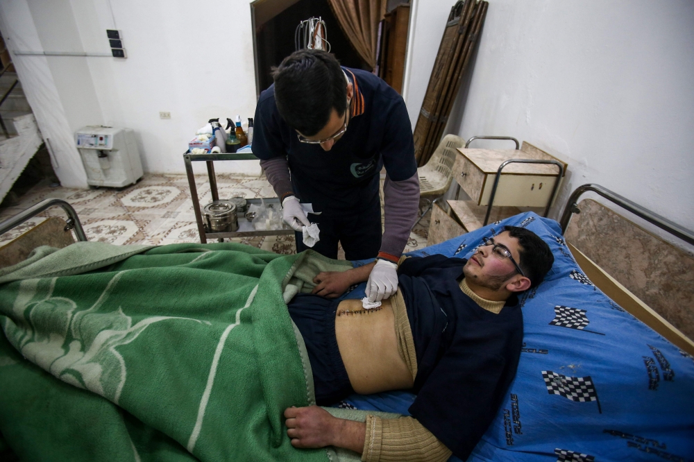 TOPSHOT - Malek Abu Jaber (below), a 20-year-old nurse, lies on a bed as a colleague changes his bandages at a makeshift clinic in the rebel-held town of Jisreen, in the besieged Eastern Ghouta region on the outskirts of the capital Damascus, on February 10, 2018, after he was injured on his way home earlier by an air strike. Eastern Ghouta, the last rebel bastion on the capital's doorstep, is home to around 400,000 besieged Syrians.  
In early February 2018, Syrian warplanes and artillery conducted an intense five-day campaign there that left around 250 civilians dead and triple that number wounded. / AFP / ABDULMONAM EASSA
