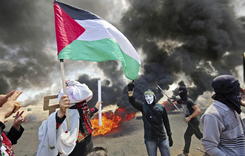 A Palestinian man holding his national flag walks in the smoke billowing from burning tyres next to a protester wearing an Anonymous mask during clashes with Israeli forces along the border with the Gaza strip east of Khan Yunis on May 14, 2018, as Palestinians protest over the inauguration of the US embassy following its controversial move to Jerusalem.  / AFP / SAID KHATIB
