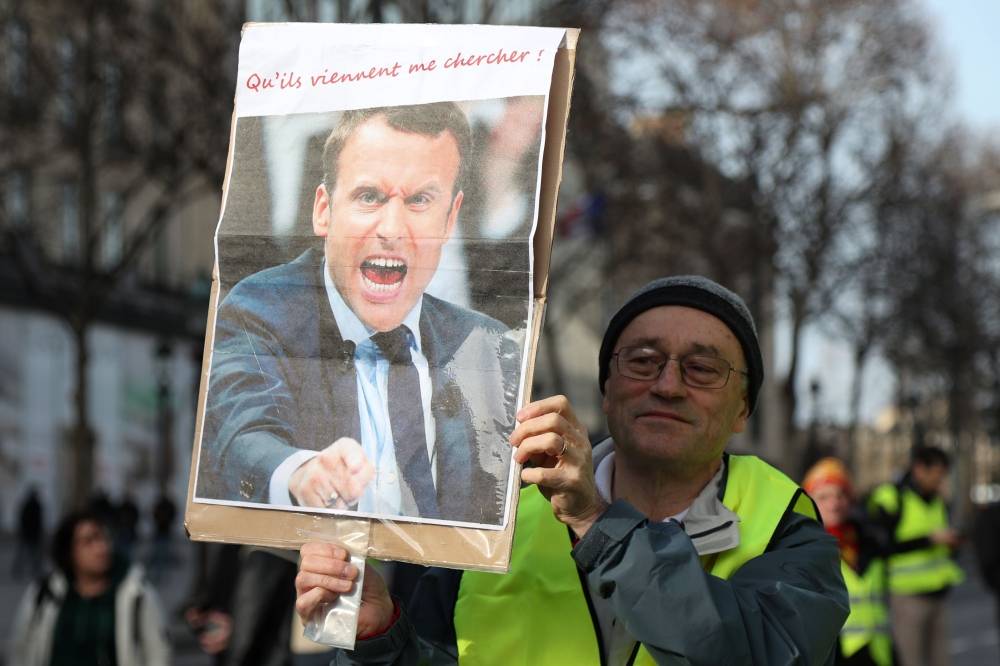 A protester holds up a placard showing French President Emmanuel Macron with the lettering 'Let them come get me' in front of the Arc de Triomphe in Paris on February 9, 2019, as 'Yellow vest' protesters (Gilets Jaunes) take to the streets for 13th consecutive Saturday. / AFP / Zakaria ABDELKAFI 