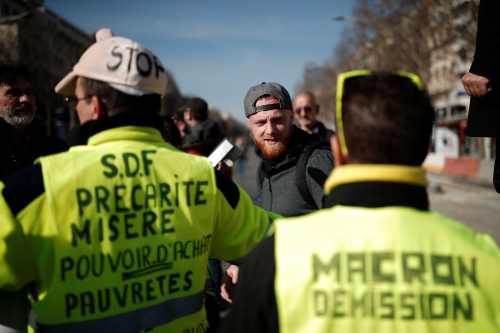 Yellow vest protester Maxime Nicolle is framed by people wearing yellow vests as they take part in a demonstration by the «yellow vests» movement in Paris, France, February 16, 2019. Messages read, «Homeless, Precarituy, Misery, Purchasing Power» and «Macron Resign». REUTERS/Benoit Tessier