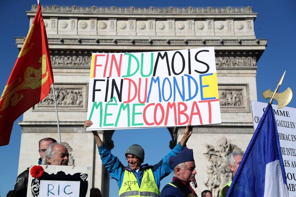 French activist Jean-Baptiste Redde, aka Voltuan, (C) holds a sign reading «End of the month, emd of the world, same fight» in front of the Arc de Triomphe during an anti-government demonstration on February 23, 2019 in Paris, as «Yellow Vest» protesters take to the streets for the 15th consecutive Saturday. The «Yellow Vest» (Gilets Jaunes) movement in France originally started as a protest about planned fuel hikes but has morphed into a mass protest against President's policies and top-down style of governing. / AFP / Zakaria ABDELKAFI 