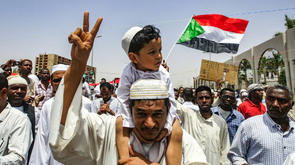 A Sudanese man carrying a child on his back flashes the victory gesture while marching with others during a rally demanding a civilian body to lead the transition to democracy, outside the army headquarters in the Sudanese capital Khartoum on April 11, 2019. Sudanese protesters angry that army commanders have taken control after removing veteran president Omar al-Bashir in a palace coup defied a night-time curfew to keep up four months of mass demonstrations today. / AFP / Ebrahim Hamid
