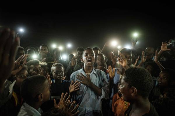 TOPSHOT - People chant slogans as a young man recites a poem, illuminated by mobile phones, before the opposition's direct dialog with people in Khartoum on June 19, 2019. People chanted slogans including «revolution» and «civil» as the young man recited a poem about revolution. / AFP / Yasuyoshi CHIBA 