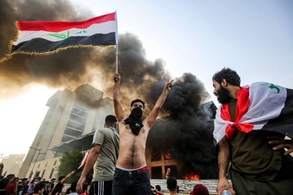 A topless masked protester flashes the victory gesture as he waves an Iraqi national flag before a burning building during a demonstration against state corruption and poor services, between the capital Baghdad's Tahrir Square and the high-security Green Zone district, on October 1, 2019. Security forces used water cannons and tear gas to disperse more than 1,000 protesters in central Baghdad. Iraq is considered the 12th most corrupt country in the world according to Transparency International. Power cuts are rampant, water shortages are common and unemployment is high, particularly among youth. / AFP / AHMAD AL-RUBAYE
