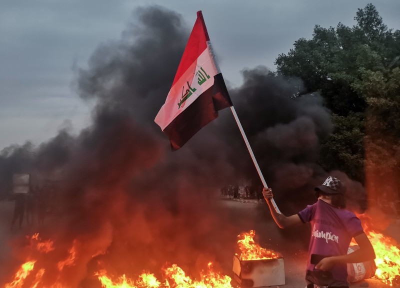 محتج عراقي يرفع علم بلاده بجوار نيران مشتعلة في مظاهرات الناصرية.